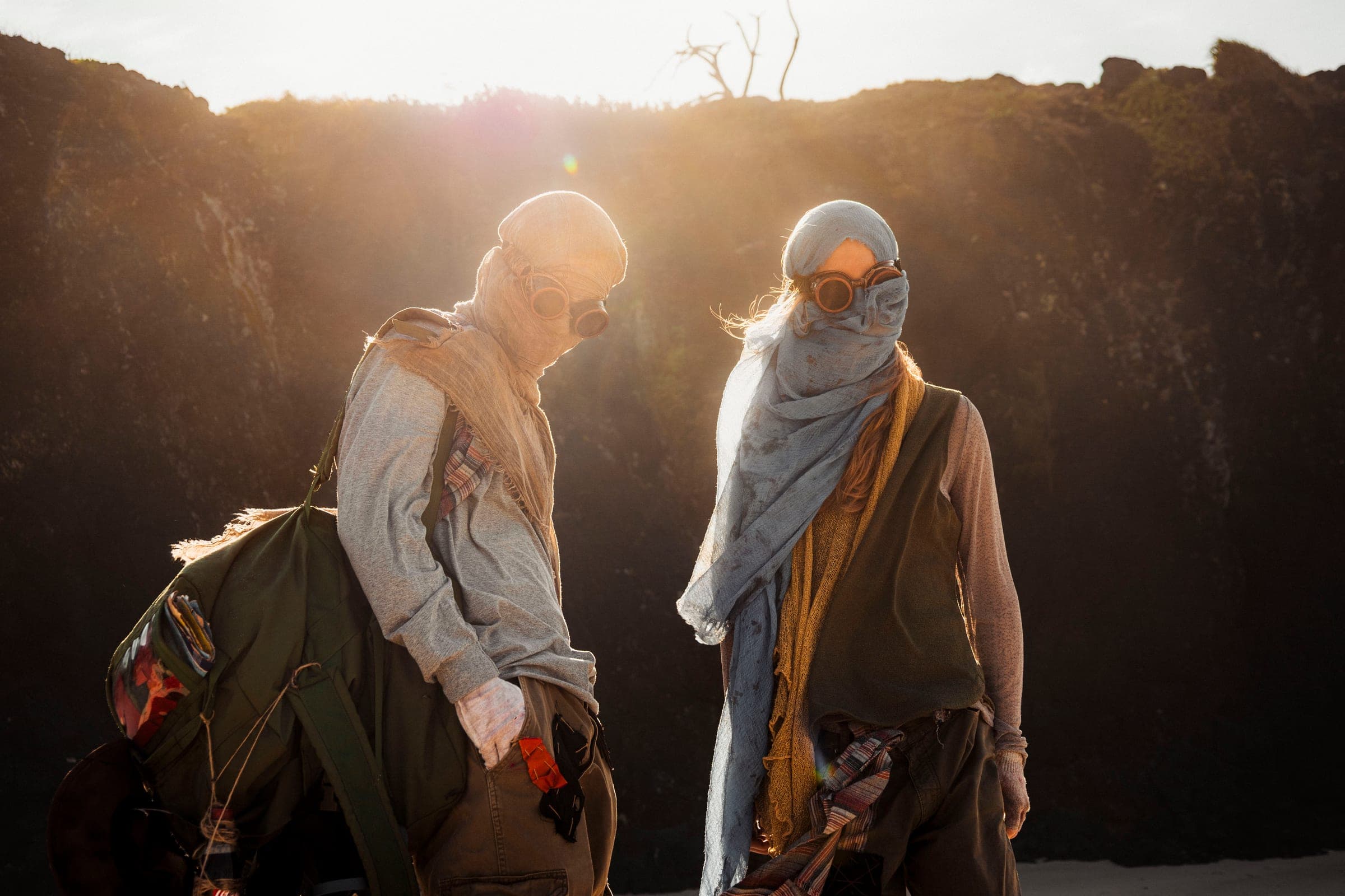 Two young people in gauze and copper goggles stand on the sand in front of a dark cliff. The sun breaks over the edge behind them, and a dead tree stands in silhouette against the sky.