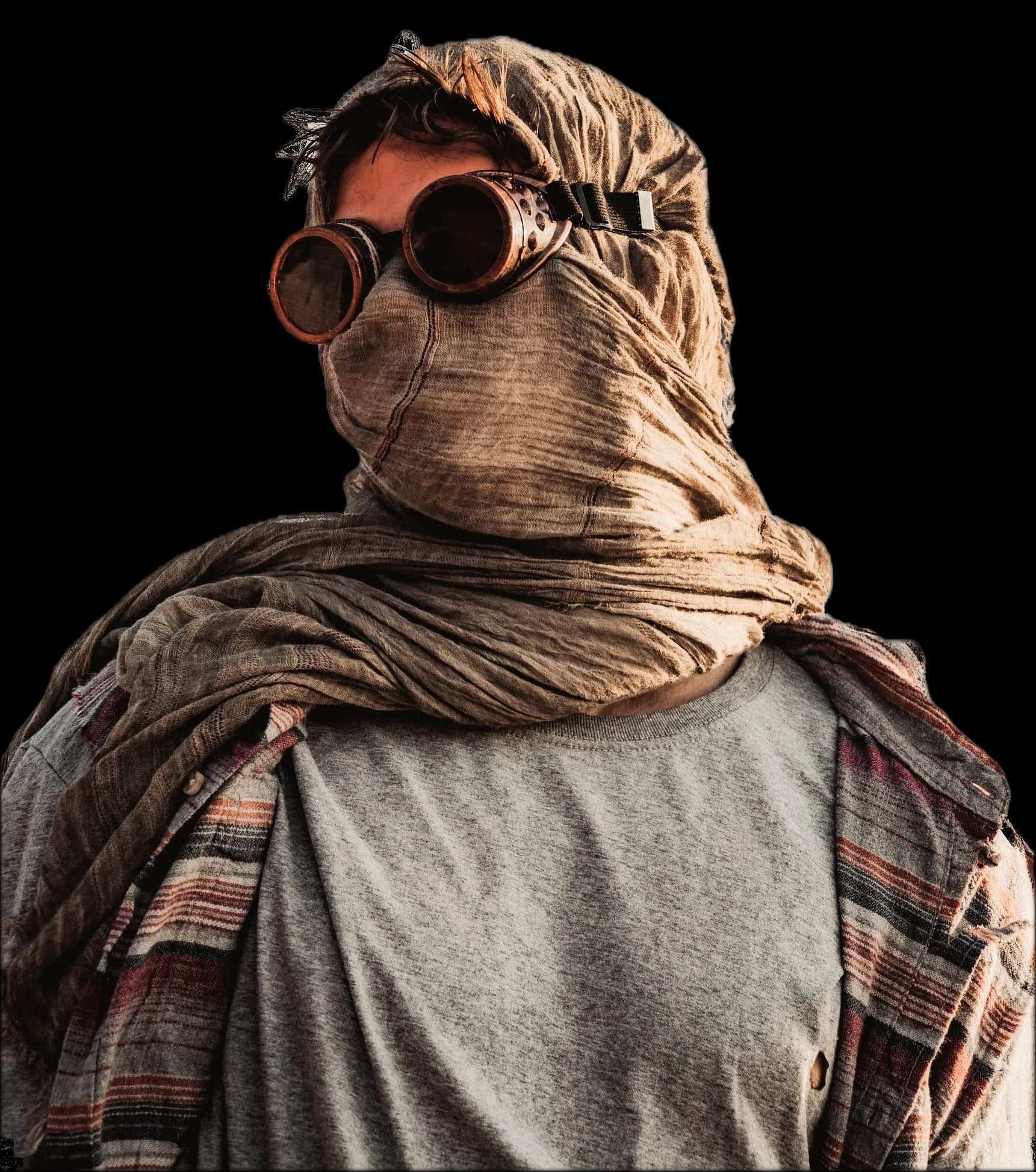 A young man seen from below against a grey sky. His head is wrapped in sand-coloured gauze, and he wears round copper goggles, a grey T-shirt and a striped scarf over his shoulders.