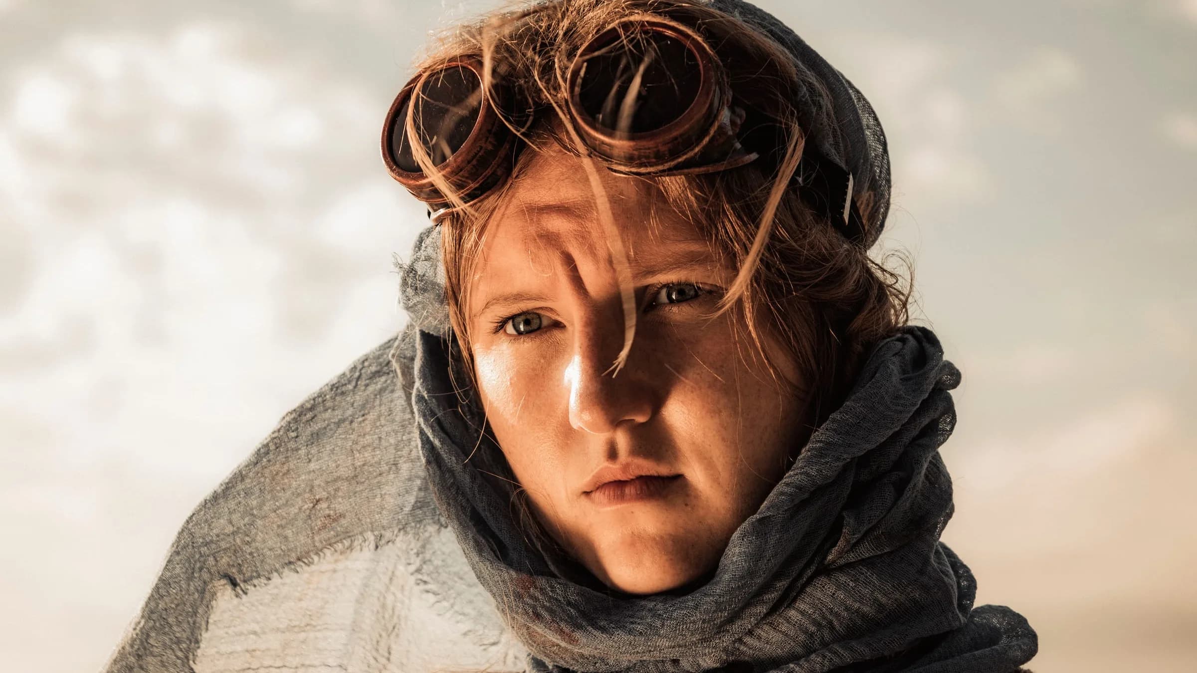 A young woman with auburn hair and freckles looks straight into the camera. Copper goggles are pushed up on her head, a blue-grey scarf is wrapped around her neck, and hard side light casts a shadow across her face.