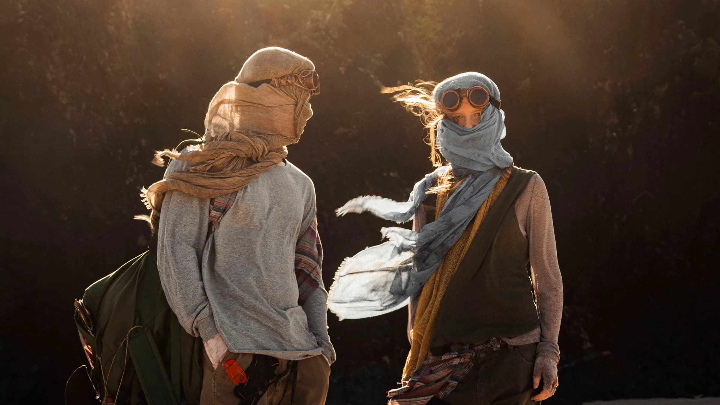 Two young people face each other in front of a dark wall as a gust of wind whips the blue scarf of the woman on the right.